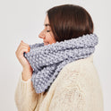 A woman is holding up the Mateusz Snood that she is wearing, it is knitted in Stormy Grey, using Stitch & Story's 100% super chunky merino wool included in the knitting kit.
