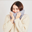A woman is cosying up in her stormy grey mateusz snood, recently knitted up from Stitch & Story's beginner friendly knitting kit.