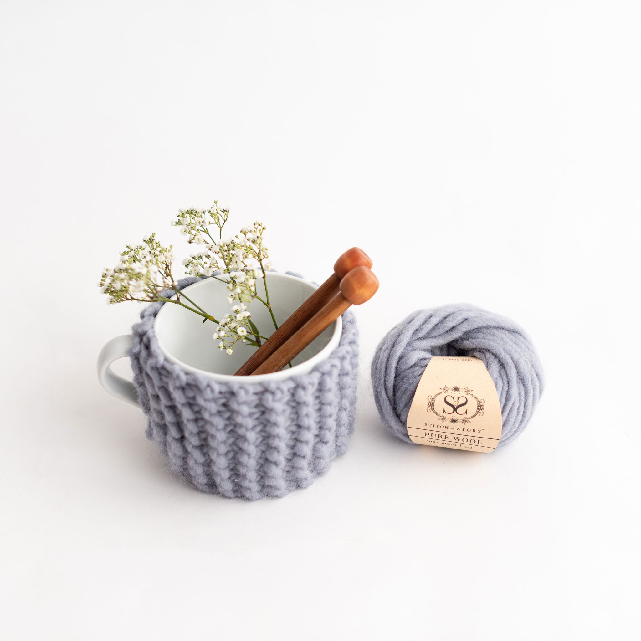 A kit content photo to show you the knitted up cup cosy in Stormy Grey around a mug, a pair of mini 10mm bamboo knitting needles and a ball of 25g Pure Wool.