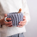 A person is holding a mug with the Stormy Grey cup cosy wrapped around the cup, and a pair of bamboo knitting needles are inside the cup.