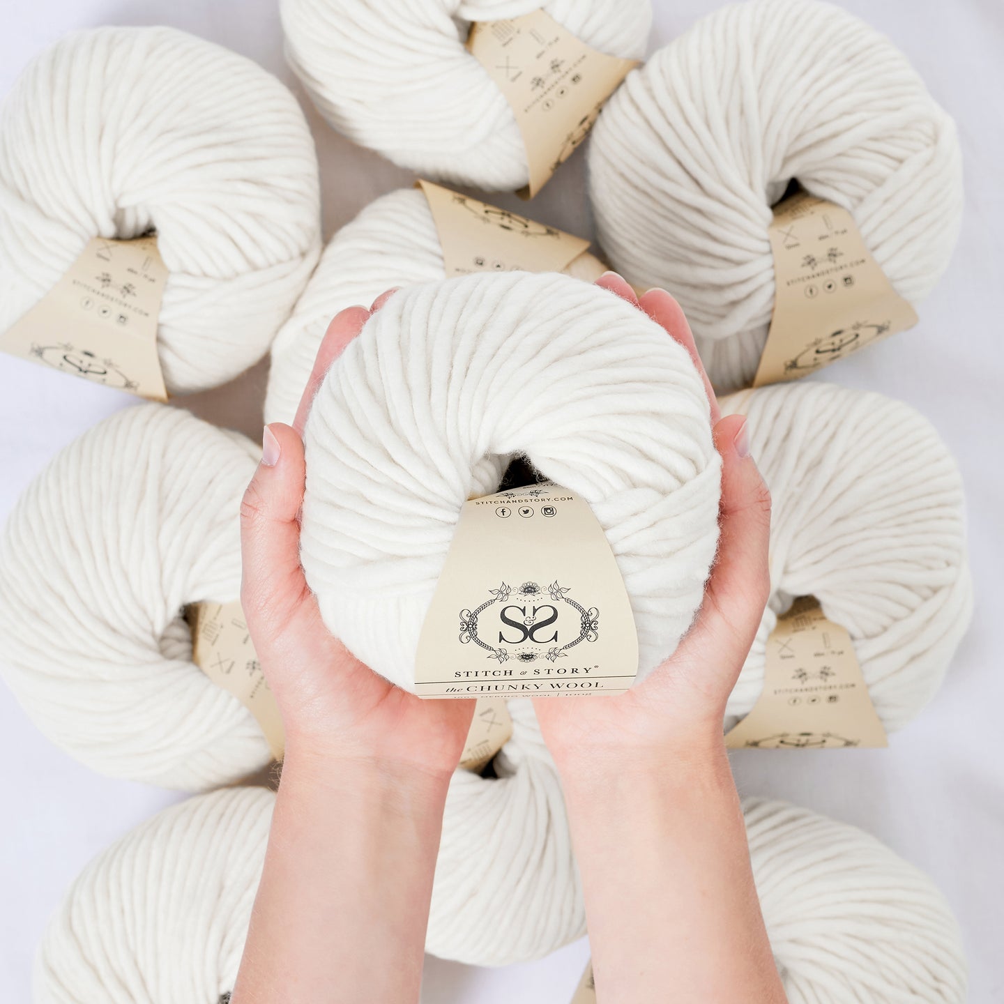 A hand is holding a ball of 100% merino wool Stitch & story The Chunky Wool in front of the camera, above another 9 balls of the same yarn. The colour shown here is pure white