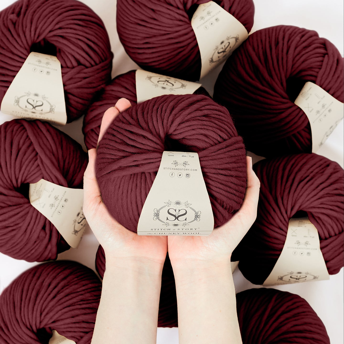 A hand is holding a ball of 100% merino wool Stitch & story The Chunky Wool in front of the camera, above another 9 balls of the same yarn. The colour shown here is sangria red