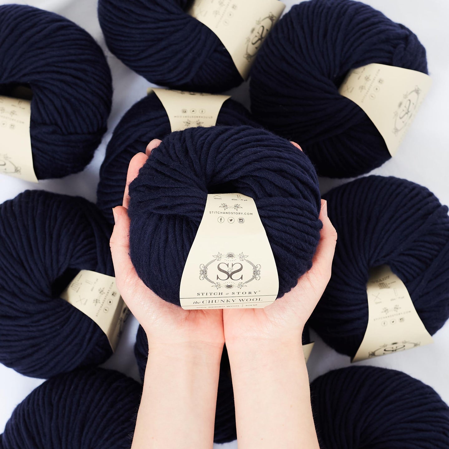 A hand is holding a ball of 100% merino wool Stitch & story The Chunky Wool in front of the camera, above another 9 balls of the same yarn. The colour shown here is silent night