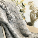 A close up of a Dove Grey Ivy Geometric blanket draped on a chair, showing the stitch definition and a tassel.
