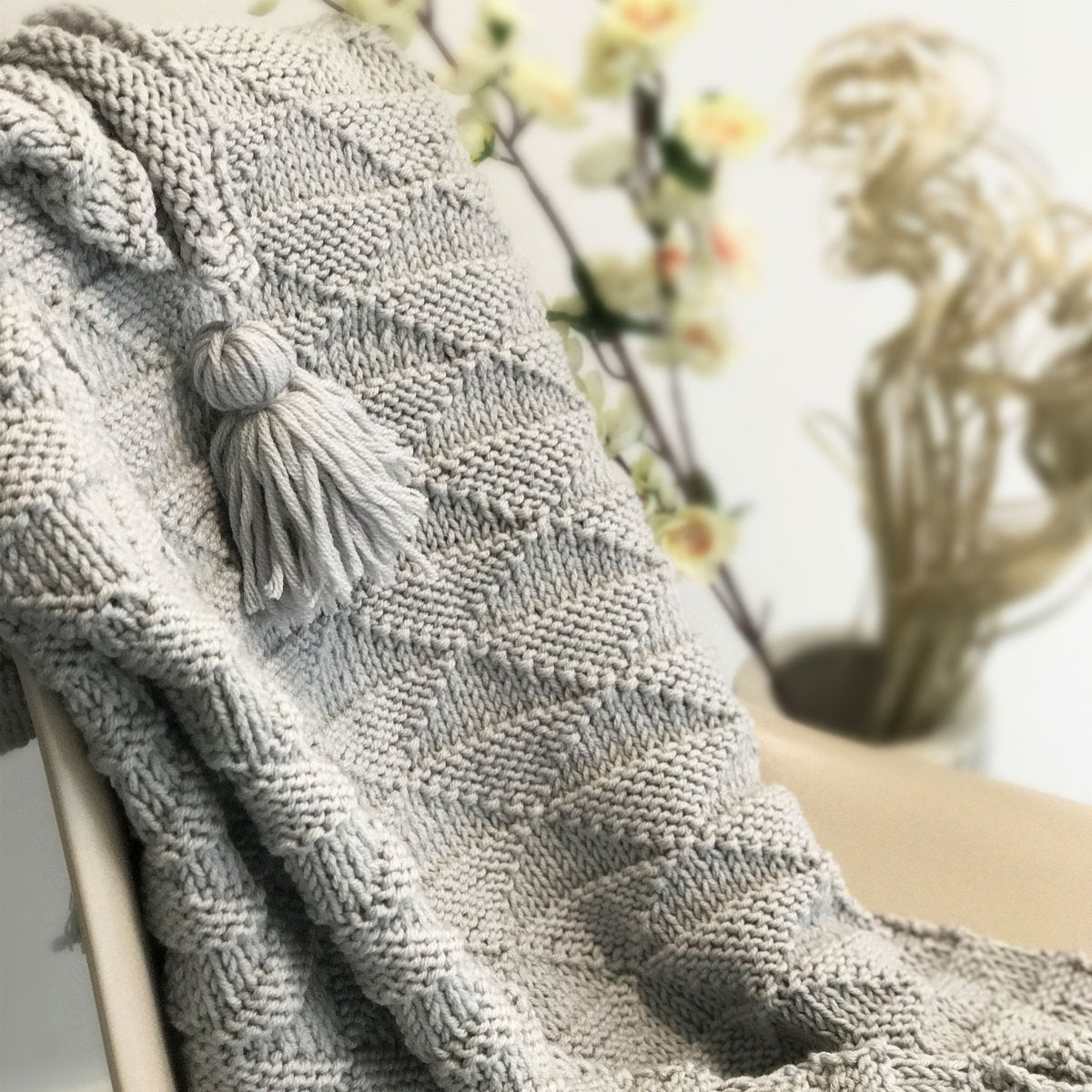 A close up of a Dove Grey Ivy Geometric blanket draped on a chair, showing the stitch definition and a tassel.