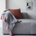 A stormy grey Chunky tassel throw with dust pink tassels is draped softly over a grey fabric sofa.